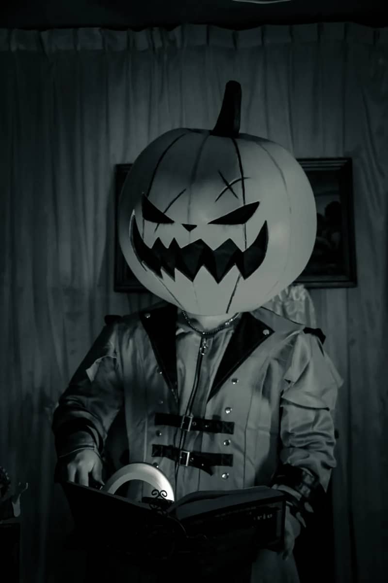 Grainy black-and-white portrait of a figure in a jack-o-lantern mask holding an open book